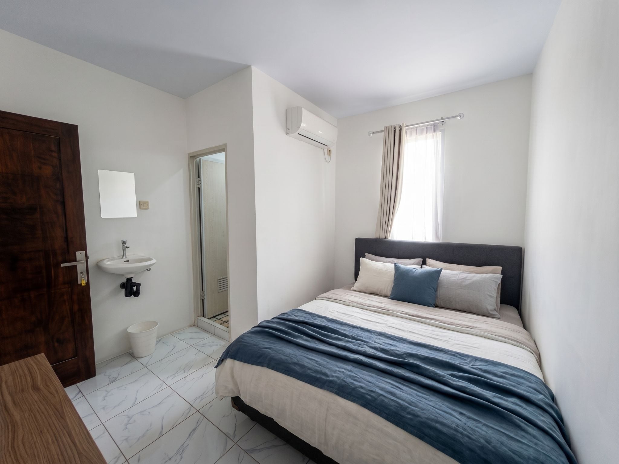 A bedroom with a bed, a bedside table, a mirror, a window with curtains, and a door.