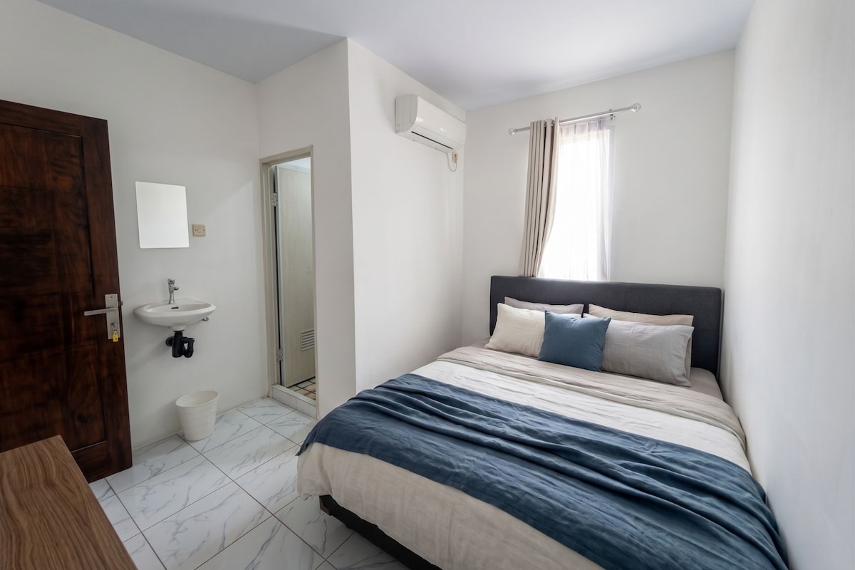 Un dormitorio con una cama, una mesita de noche, un espejo, una ventana con cortinas y una puerta.