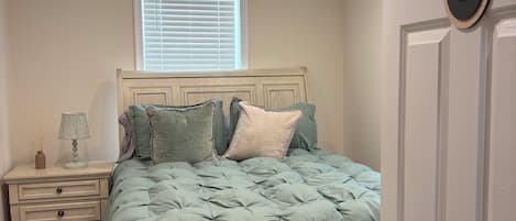Une chambre à coucher avec un lit, une table de chevet, une lampe, une fenêtre avec des stores et une porte portant un numéro.