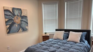 Ein Schlafzimmer mit einem großen Bett, einem Nachttisch, einem Wandbild und einem Fenster mit Jalousien.