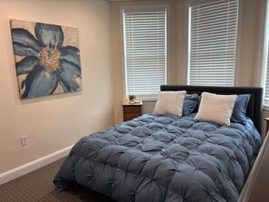 Quarto com cama grande, mesa de cabeceira, quadro na parede e janela com persianas.