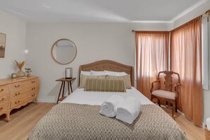 A neatly arranged bedroom with a bed, a wooden dresser, a chair, and a bedside table. - The Corner Saloon with desert and mountain views (Twentynine Palms)