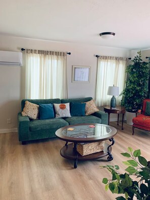 A living room with a green sofa, a glass coffee table, a red chair, a potted plant, and a wall-mounted air conditioner. - The Corner Saloon with desert and mountain views (Twentynine Palms)