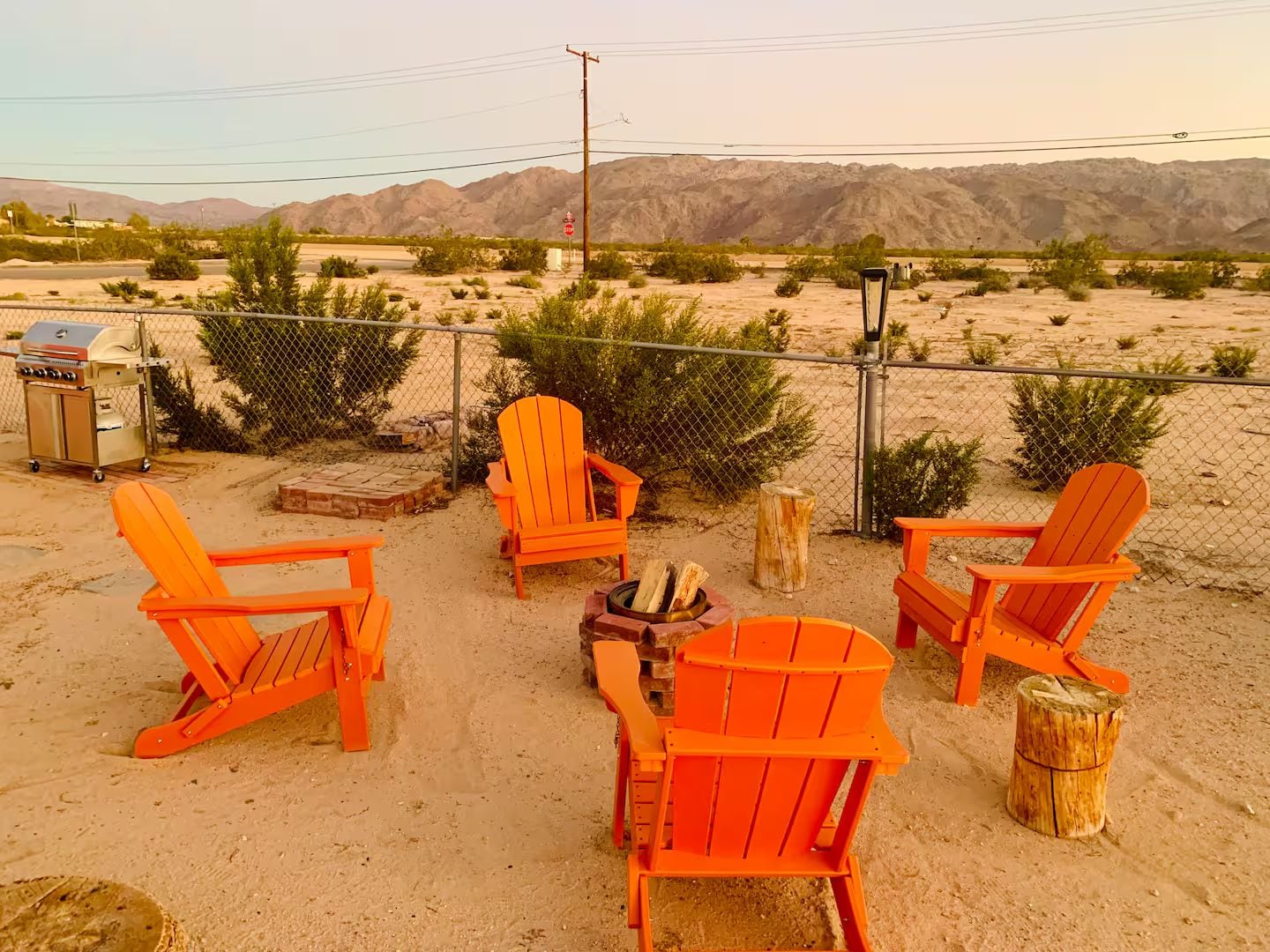 An outdoor seating area with orange chairs and a grill, surrounded by a chain-link fence with desert vegetation and mountains in the background.