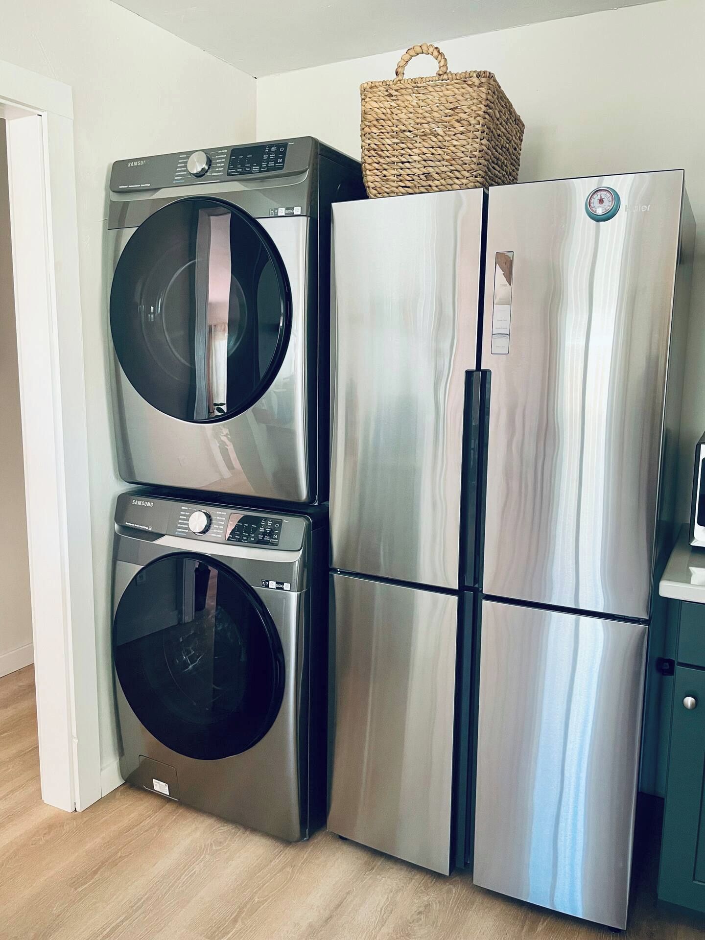 A stainless steel refrigerator and a washing machine stacked in a corner with a woven basket on top.