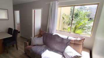 Een woonkamer met een bank, een eettafel met stoelen, een raam met gordijnen en uitzicht op groen buiten.