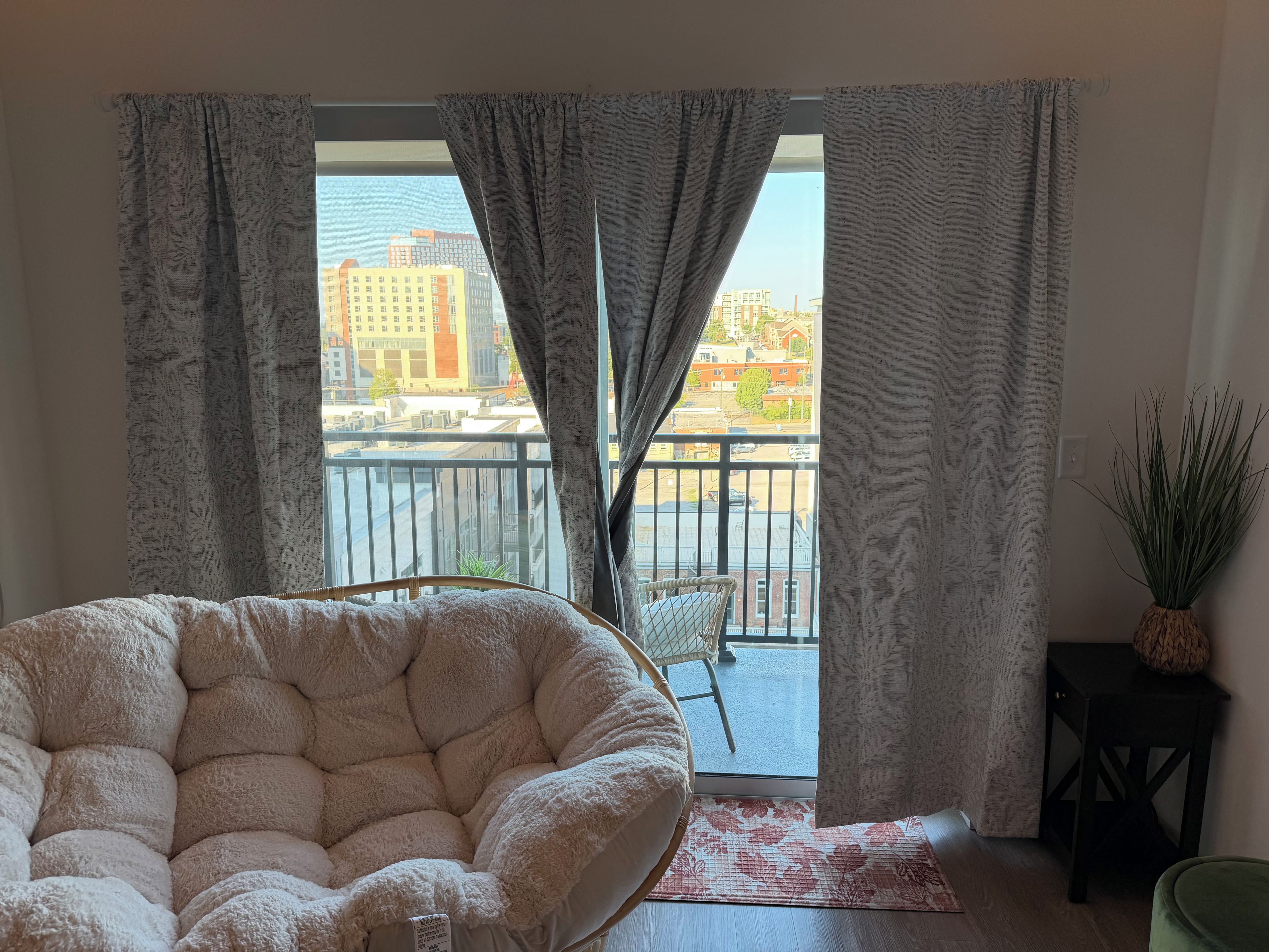 Une pièce comprenant un grand fauteuil moelleux, une petite table avec une plante et une vue sur un balcon donnant sur des immeubles.