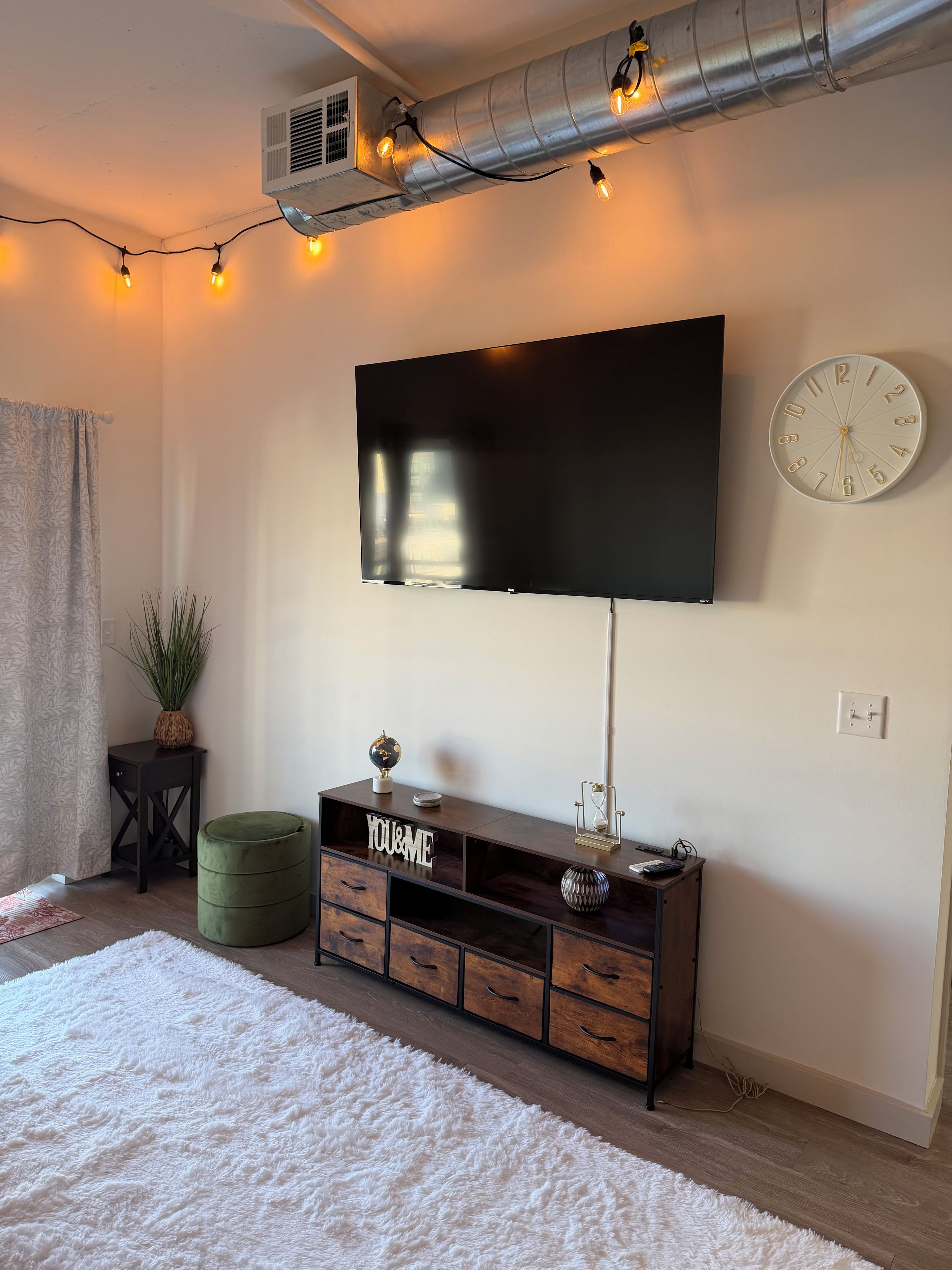 Een moderne woonkamer met een flatscreen televisie aan de muur, een houten kast met lades, een groene poef, een zacht wit vloerkleed en een klok.