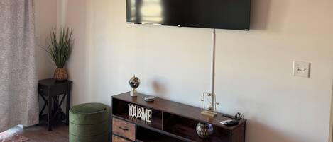 Un salon moderne avec un téléviseur à écran plat fixé au mur, un meuble en bois avec des tiroirs, un pouf vert, un tapis blanc et moelleux, et une horloge.