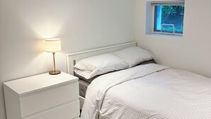 A bedroom with a bed, bedside table, lamp, and a window with a view of greenery. - Bright + Spacious Basement Suite by the Beach (VANCOUVER)