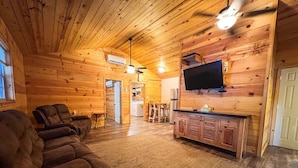 Interior de cabana de madeira com sofá, televisão e área de cozinha.