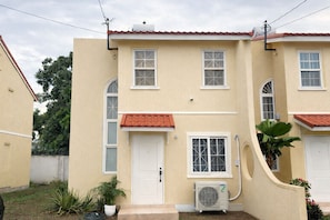 A two-story building with a red-tiled roof, a central door, and an air conditioning unit. - Jacar Purple Blossom a cozy 2-bedroom townhome (Spanish Town)