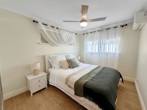 Un dormitorio con cama, mesita de noche, lámpara, ventilador de techo y ventana con cortinas.