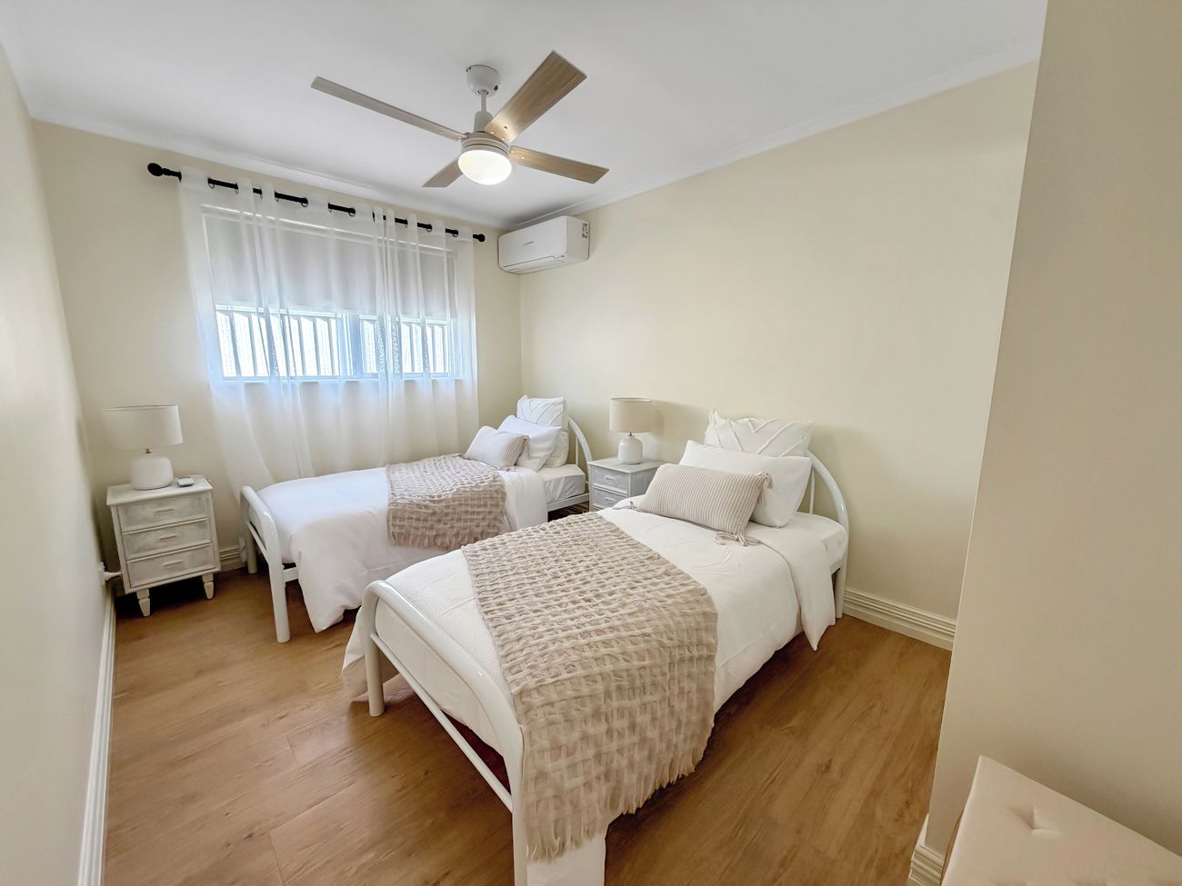 Un dormitorio con dos camas, ventilador de techo, unidad de aire acondicionado, mesita de noche con lámpara y ventana con cortinas.
