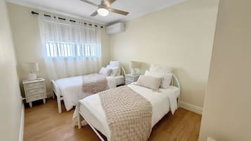 Un dormitorio con dos camas, ventilador de techo, unidad de aire acondicionado, mesita de noche con lámpara y ventana con cortinas.