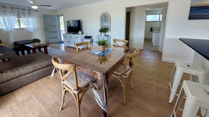 Amplia sala de estar con una mesa de comedor de madera, sillas acolchadas, un ventilador de techo y vista hacia la cocina.