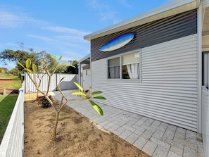 Casa móvel térrea com fachada de metal corrugado, uma janela com cortinas e um pequeno jardim com árvores jovens.