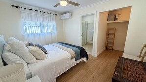 Un dormitorio con cama, ventilador de techo, ventana con cortinas, piso de madera y una pequeña mesa con silla.
