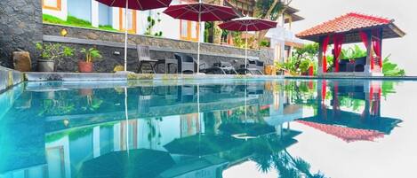 Una piscina con agua cristalina, rodeada de sillones de descanso y sombrillas, y un edificio con fachada de piedra y detalles en rojo.