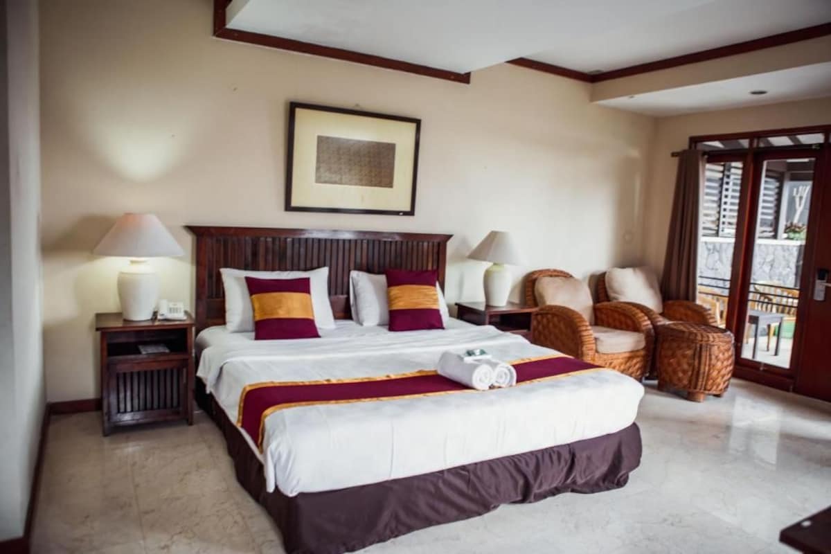 A hotel room with a large bed, two bedside tables with lamps, a framed picture on the wall, and wicker chairs by the window.