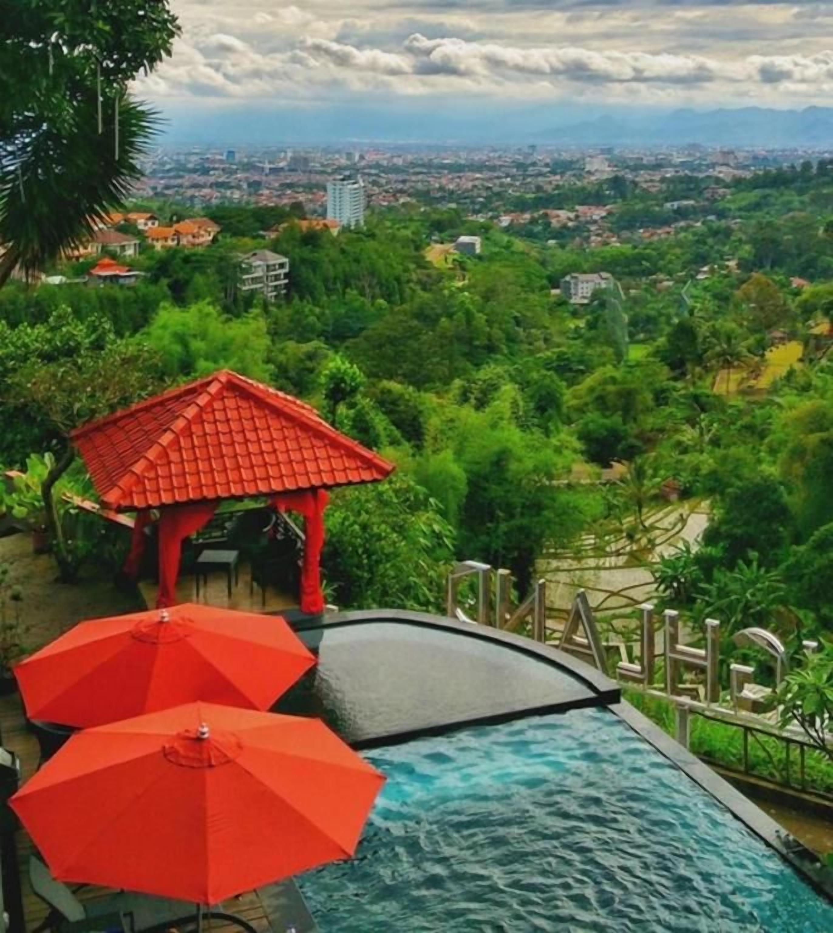 A swimming pool with a red umbrella and a gazebo, surrounded by lush greenery and a cityscape view.