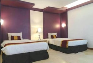 Habitación de hotel con dos camas, cada una con una colcha blanca y una almohada a rayas rojas y amarillas. Entre las camas hay una mesita de noche con una lámpara.