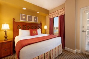 Quarto de hotel com uma cama grande, duas luminárias laterais, uma janela com cortinas vermelhas e uma porta com persianas.