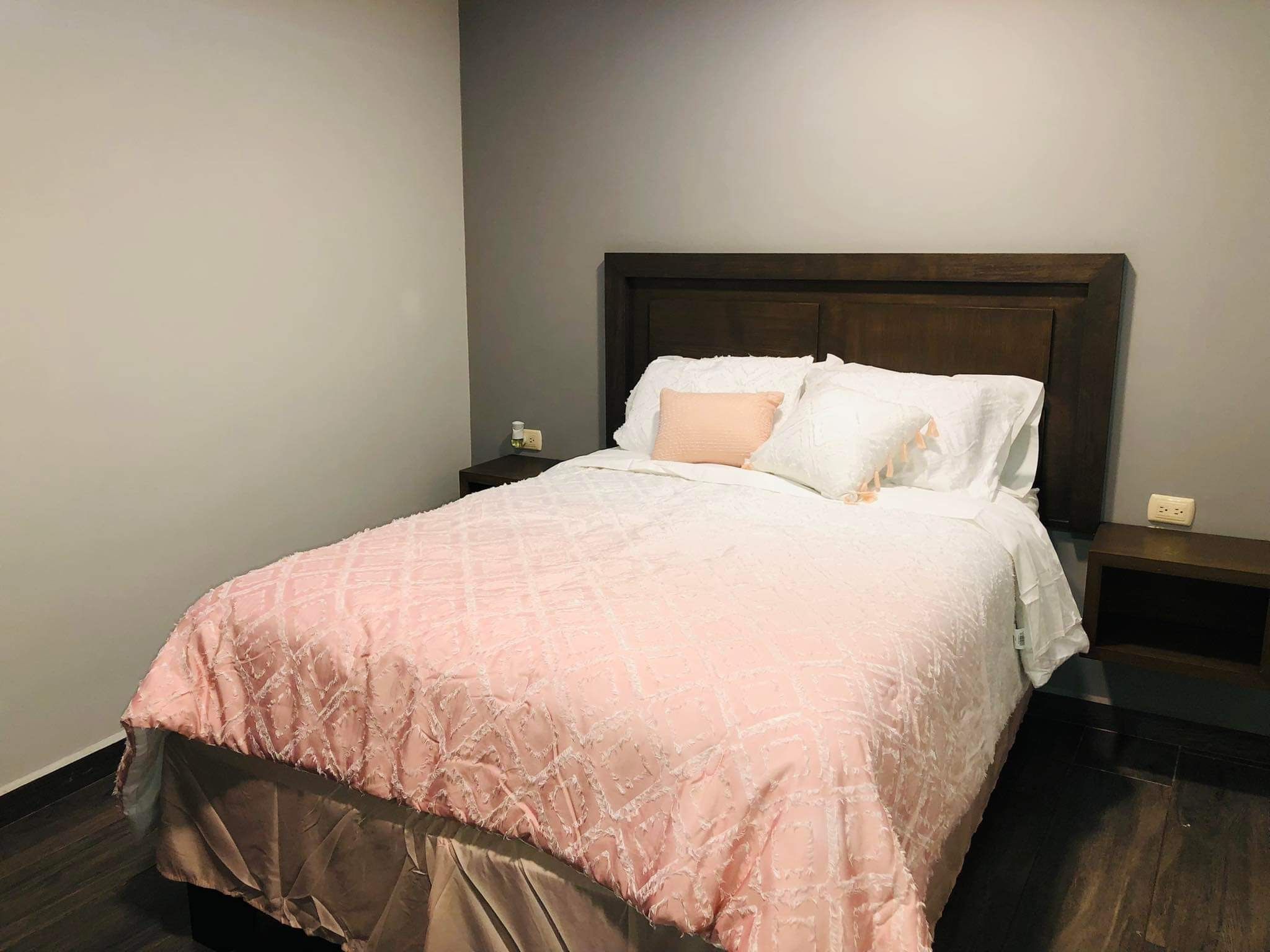 Un llit ben fet amb un edredó rosa i almoades blanques en una habitació d'hotel amb terres de fusta fosca i un capçal també fosc.