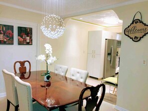 A dining area with a wooden table, white upholstered chairs, a hanging chandelier, and a 'Welcome' sign.