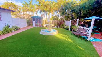 A well-maintained lawn with a central fountain, surrounded by a fence and palm trees.