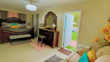 A bedroom with a bed, a dresser, a vanity, and a door leading to a balcony with a view of greenery.