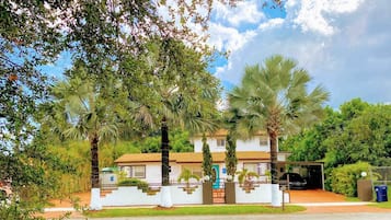 A two-story house with a white facade, a brown roof, and a front yard with palm trees.