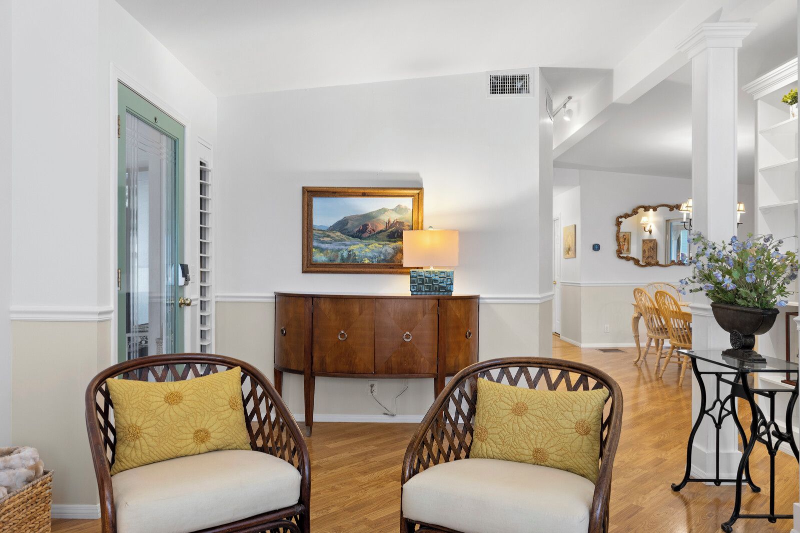 A spacious living area with a wooden cabinet, two wicker chairs with yellow cushions, a dining table with chairs, and a painting on the wall.