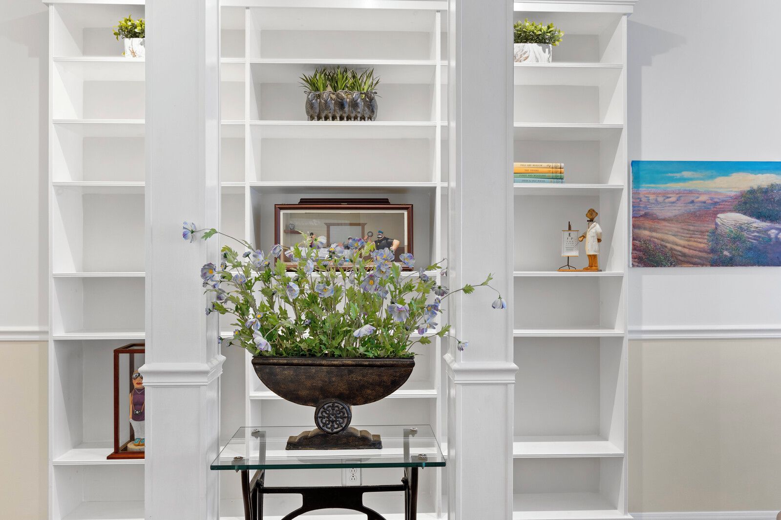 A room with white shelving units, a glass table with a decorative bowl, and a framed picture on the wall.