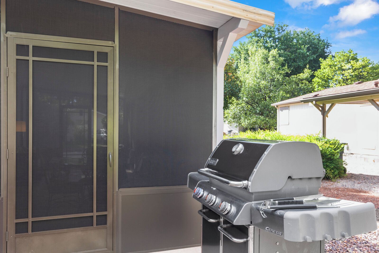 A barbecue grill with a control panel, a sliding glass door, and a covered outdoor area with a view of greenery.