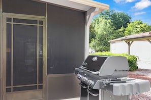 A barbecue grill with a control panel, a sliding glass door, and a covered outdoor area with a view of greenery.