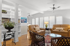A living room with two leather sofas, a glass coffee table, a black metal side table, a wicker chair, a floor lamp, and a ceiling fan.