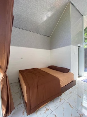 A single bed with a brown bedspread in a room with a white and gray patterned ceiling, a glass door leading outside, and a tiled floor.