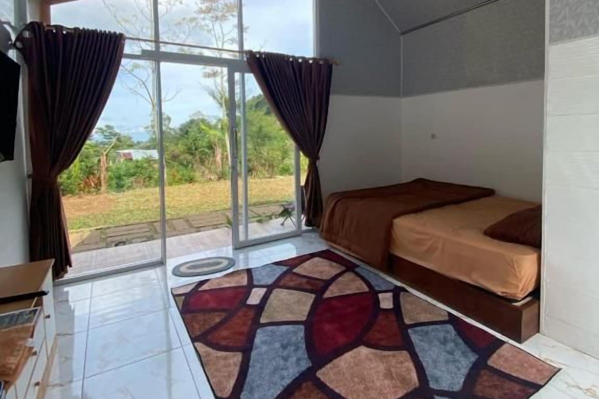 A room with a bed, a window with curtains, a patterned rug, and a view of greenery outside.