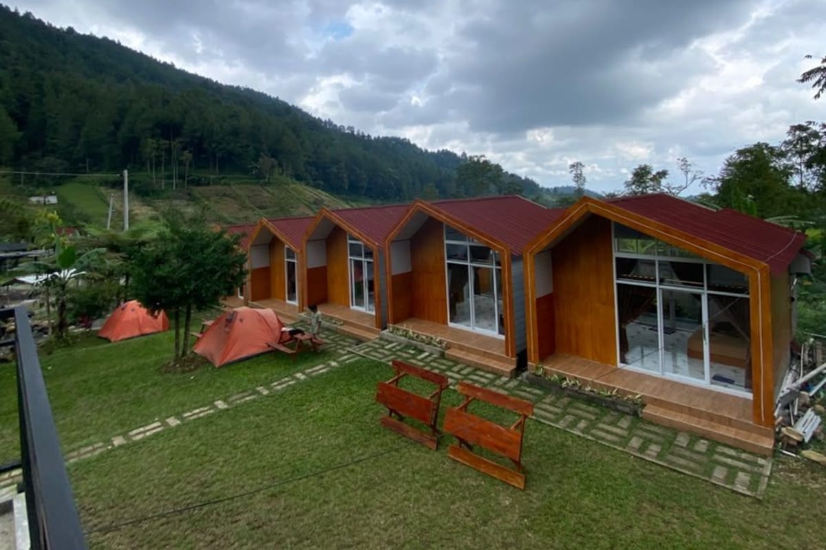Una hilera de cabañas de madera con techos rojos, un patio cubierto de césped y montañas al fondo.