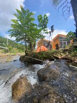 Entorno natural con un arroyo, rocas y un edificio con amplios ventanales.