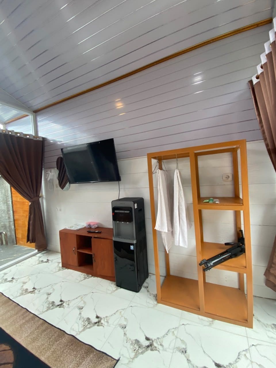 A room with a flat-screen TV mounted on the wall, a wooden cabinet, a water dispenser, and a shelf with hanging items.