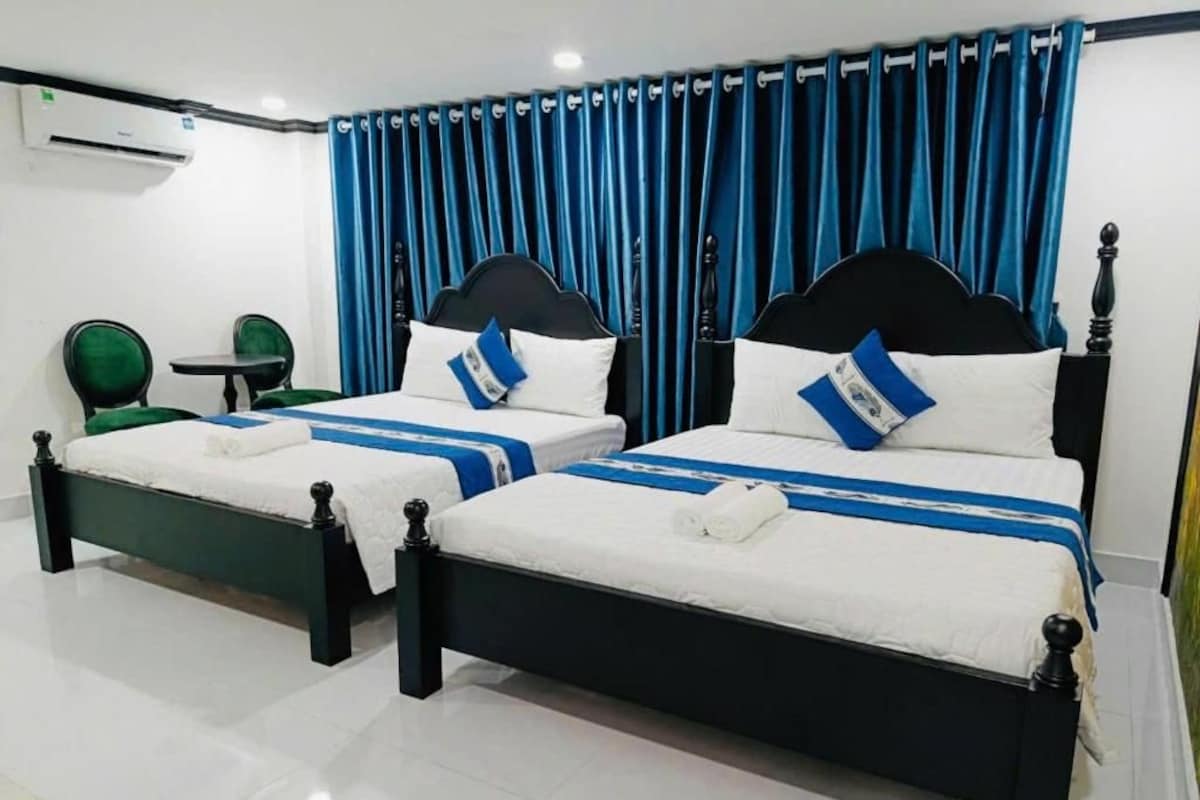 Two beds with black frames and white bedding, a small table, and green chairs in a room with blue curtains.