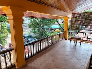 A covered balcony with a tiled floor, a wooden railing, and a view of the street and greenery outside.