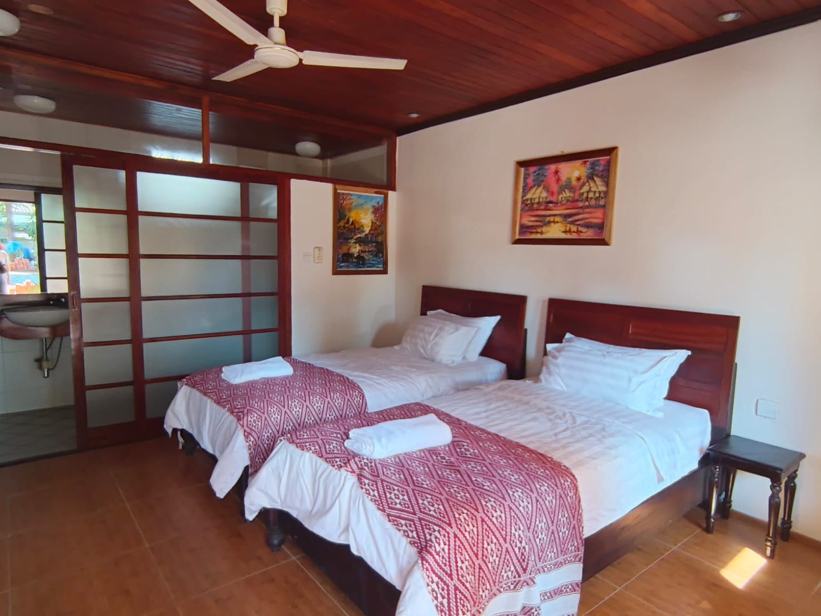 A room with two beds, a wooden ceiling, a wall-mounted painting, and a window with a view.
