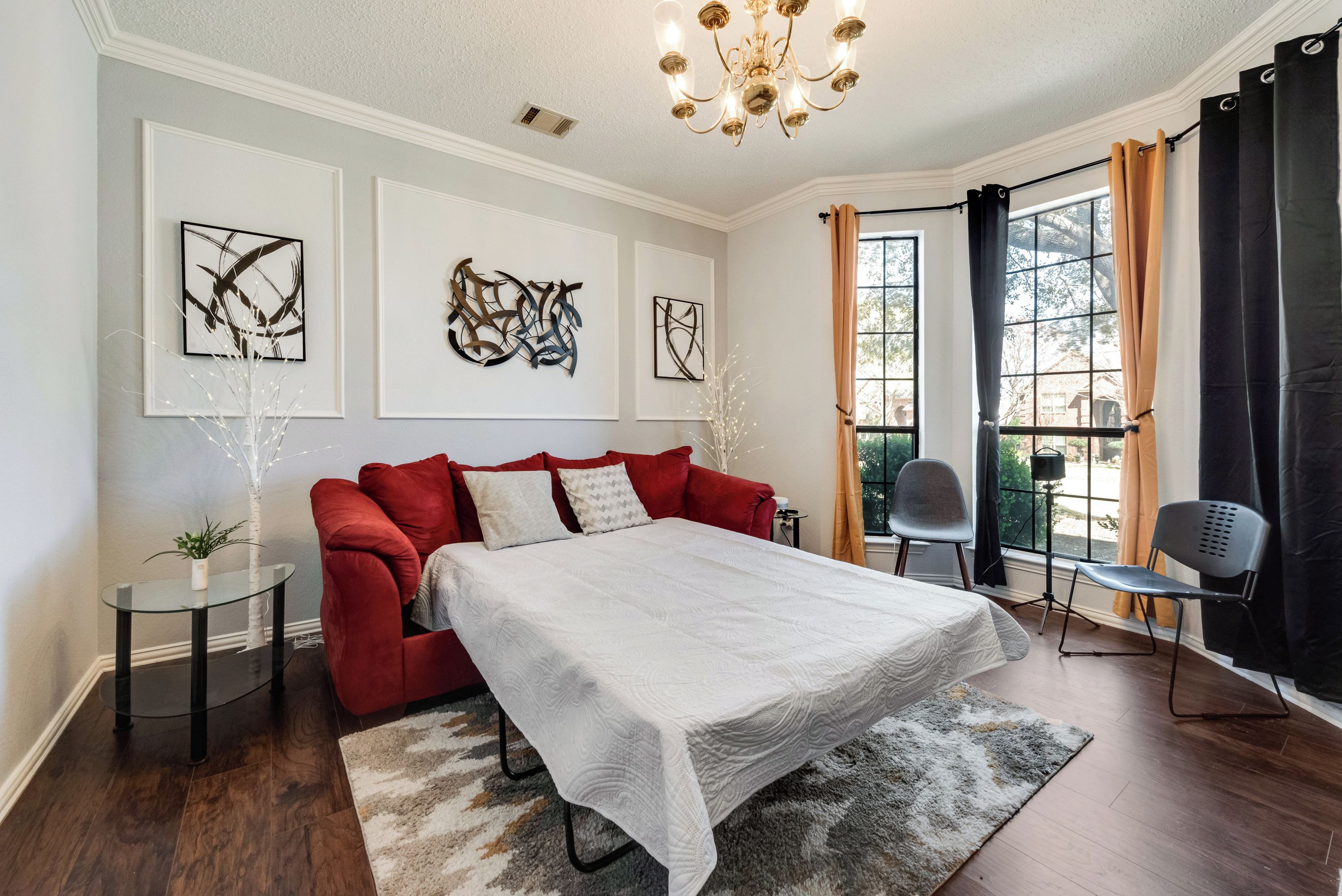 Une chambre avec un lit, un canapé rouge, une petite table et deux chaises. Les murs sont ornés d’œuvres d’art abstraites et un lustre est suspendu au plafond.