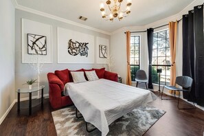 Une chambre avec un lit, un canapé rouge, une petite table et deux chaises. Les murs sont ornés d’œuvres d’art abstraites et un lustre est suspendu au plafond.