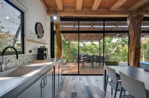 A modern kitchen with a large window, a dining area with a wooden deck, and a view of lush greenery. - Tropical Lodge 
Hidden Hideaway-House (Playa Grande)