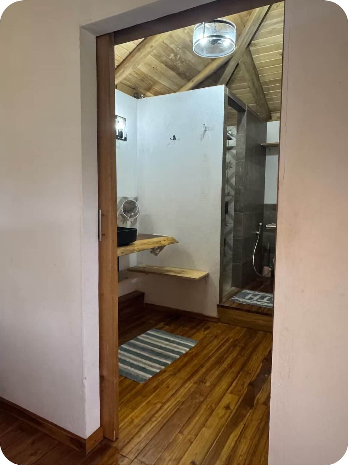 Een badkamer met een douche, wastafel en spiegel. Er ligt een houten vloer en er is een plafondventilator.