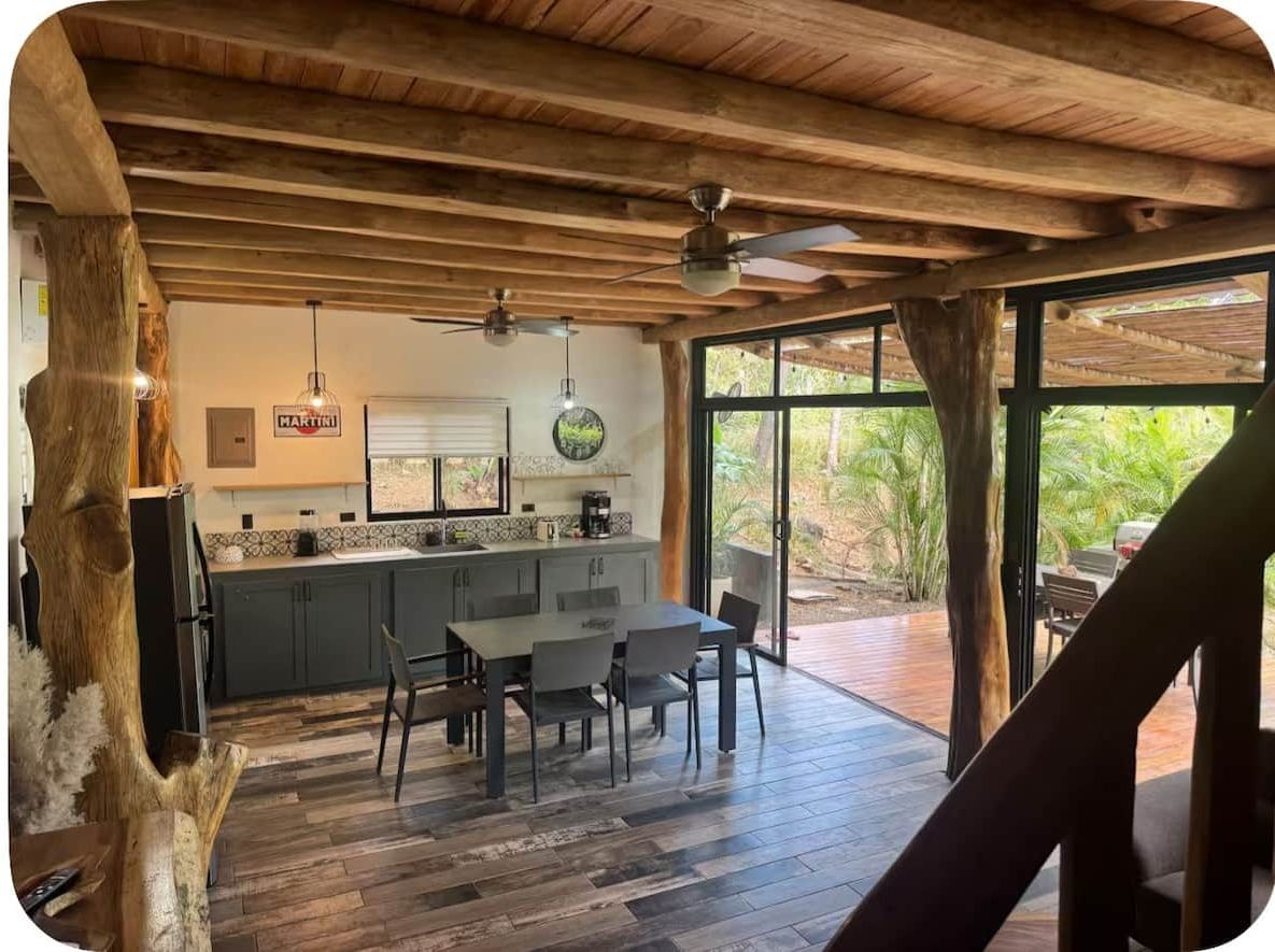 A modern kitchen with a wooden ceiling, a dining area with a table and chairs, and a large window with a view of greenery.